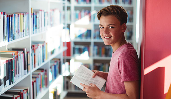 Image of a high school student reading a book while standing against a wall with stacks of book shelves around him