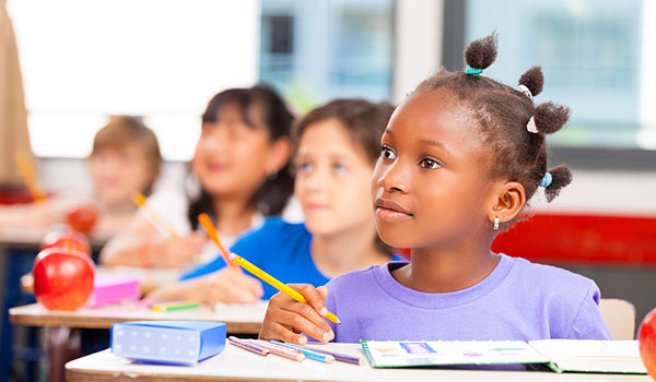 Image of an elementary student sitting at a desk looking up at a teacher with pencil in hand with three classmates blurred doing the same in the background
