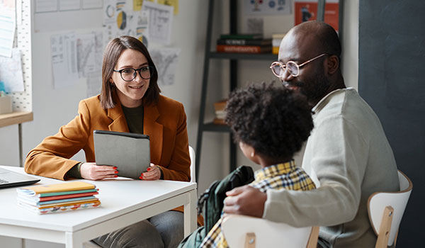 Image of a female teacher wearing glasses sitting at her desk holding a table while talking to a father and his child seated in chairs