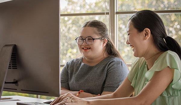 Image of a student with down syndrome wearing glasses and smiling next to a teacher as they look at a computer monitor in class